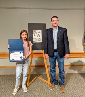 Mary Guzynski and Montana Attorney General Austin Knudsen pictured with Mary's certificate for winning the National Missing Children's Poster Contest. Her winning poster is on an easel between them.